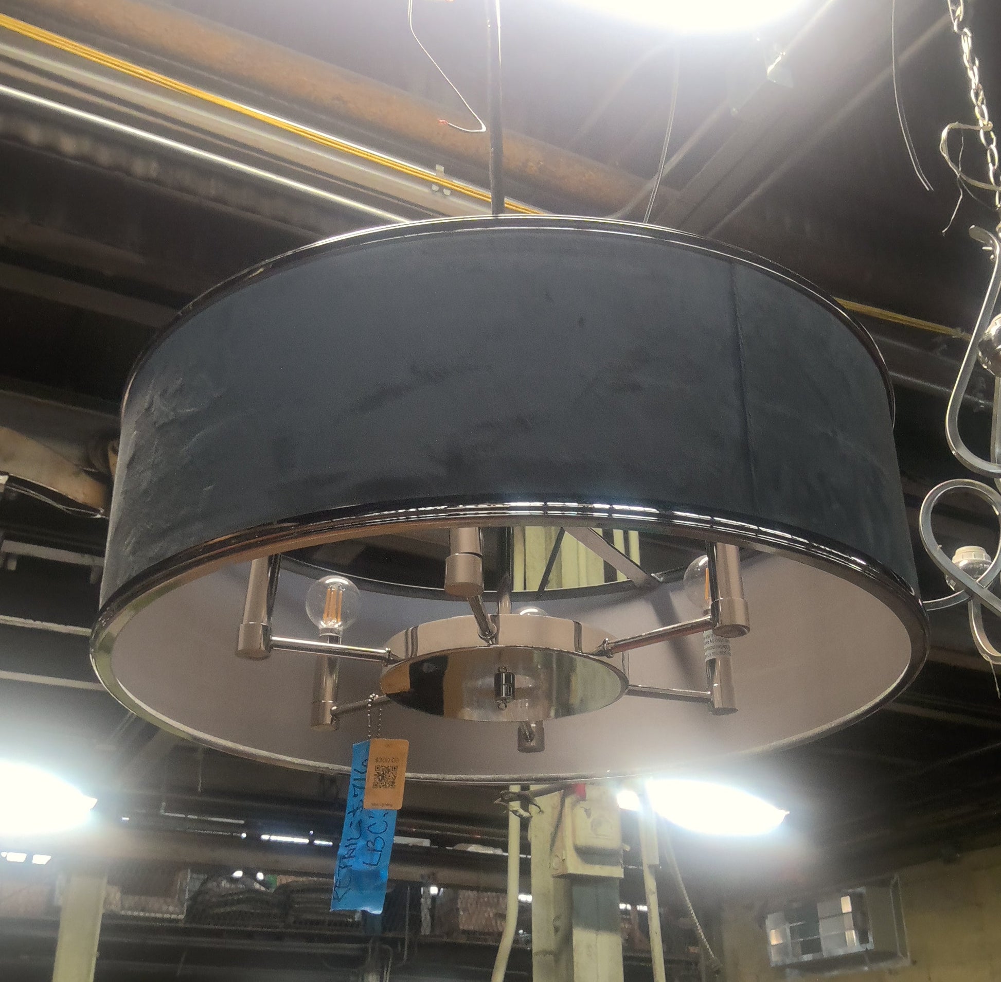 A large drum-shaped pendant light with a dark charcoal gray fabric shade and a silver metal frame, hanging in what appears to be a warehouse or furniture store.