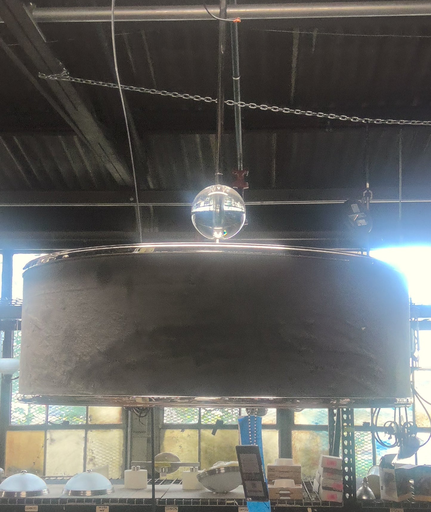 A large industrial pendant light with a wide, dark gray cylindrical shade and a clear glass globe bulb, hanging in a workshop or warehouse setting.