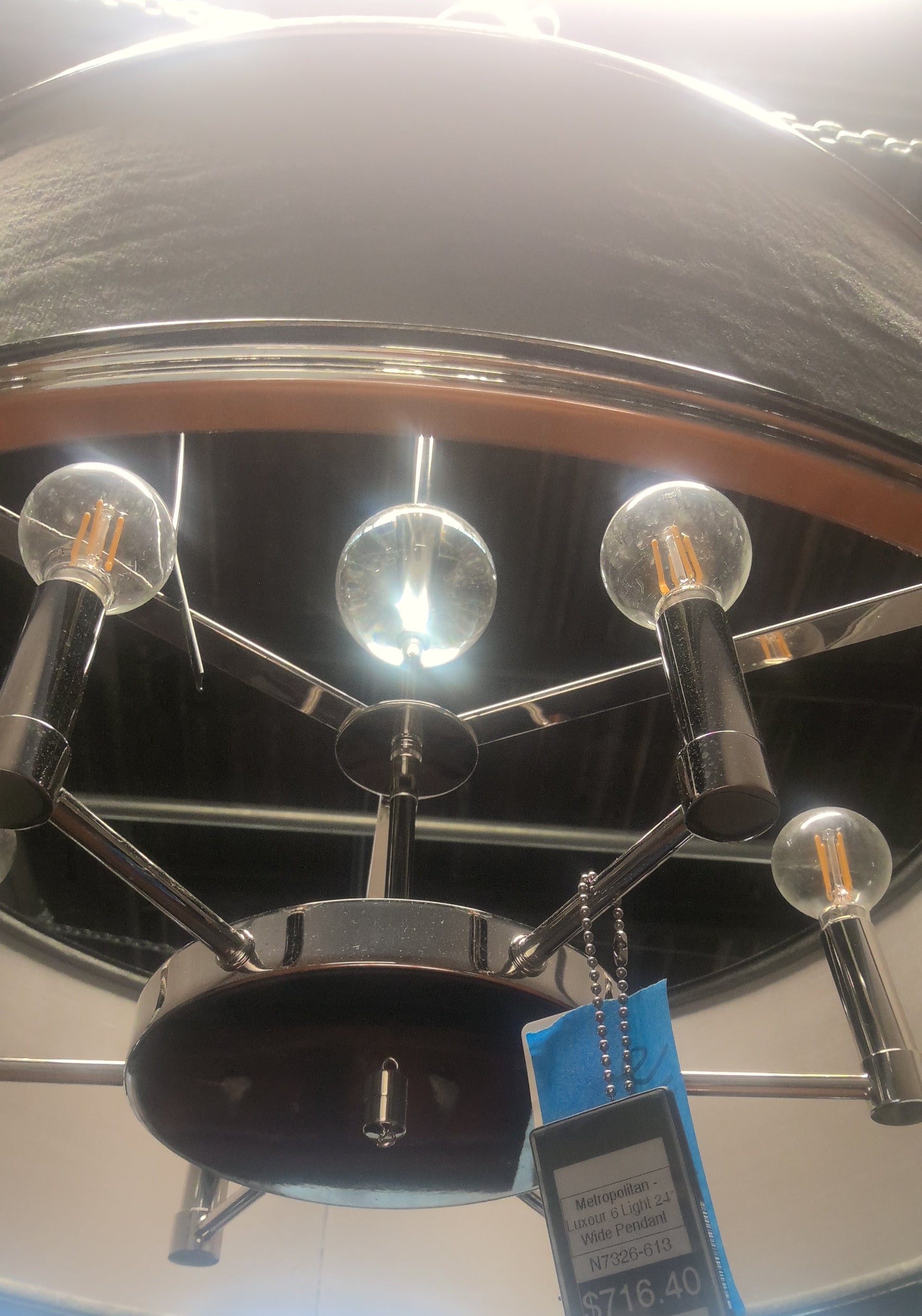 A modern multi-light chandelier with a polished chrome finish and clear glass globe bulbs, shown hanging from a ceiling in a store. A blue retail price tag is attached to the fixture.