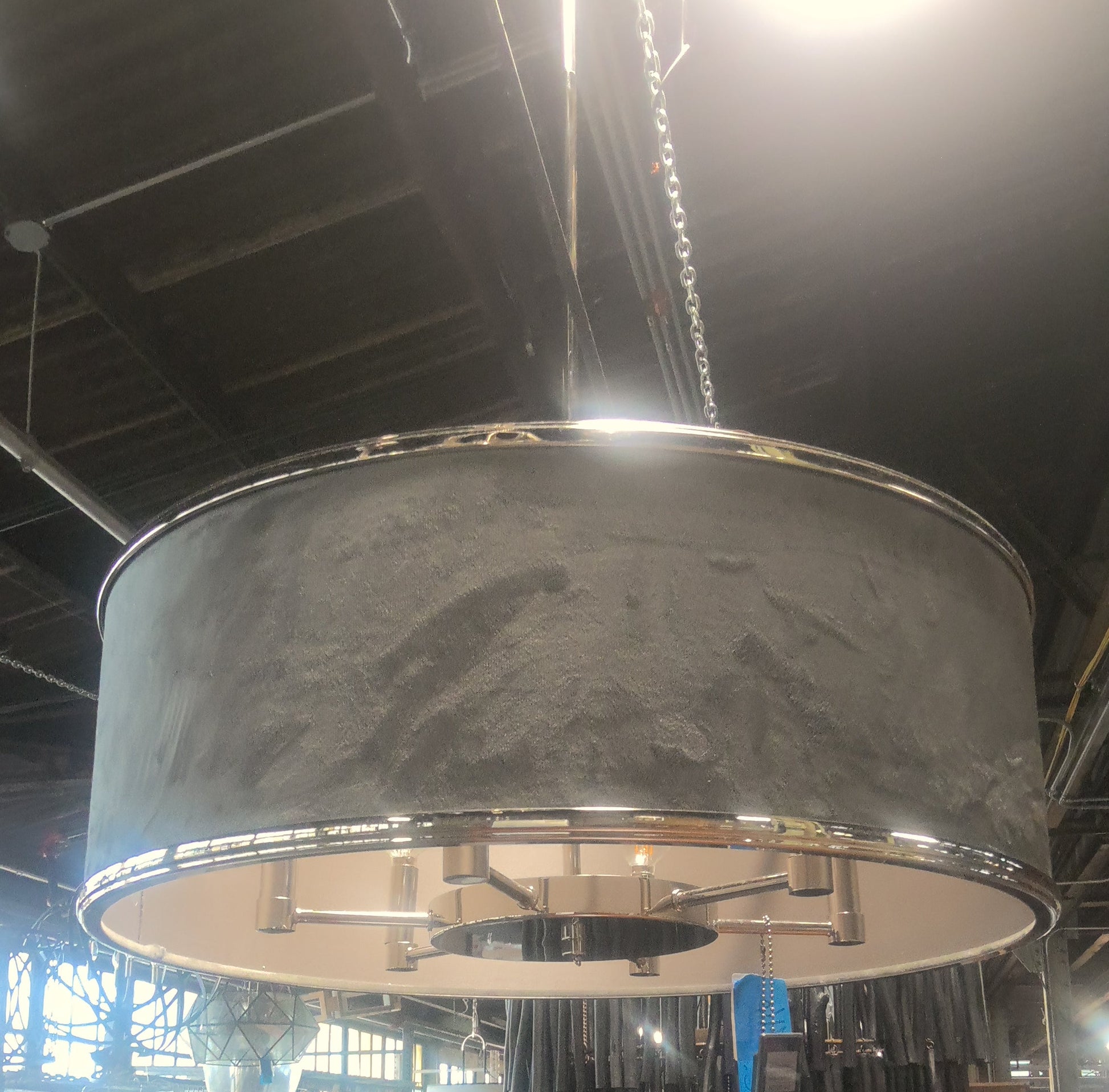 A large industrial drum pendant light with a textured gray fabric shade and polished silver metal trim, hanging in a furniture store showroom.
