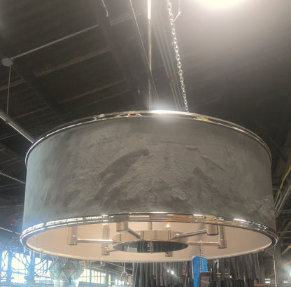 A large industrial drum pendant light with a textured gray fabric shade and polished silver metal trim, hanging in a furniture store showroom.