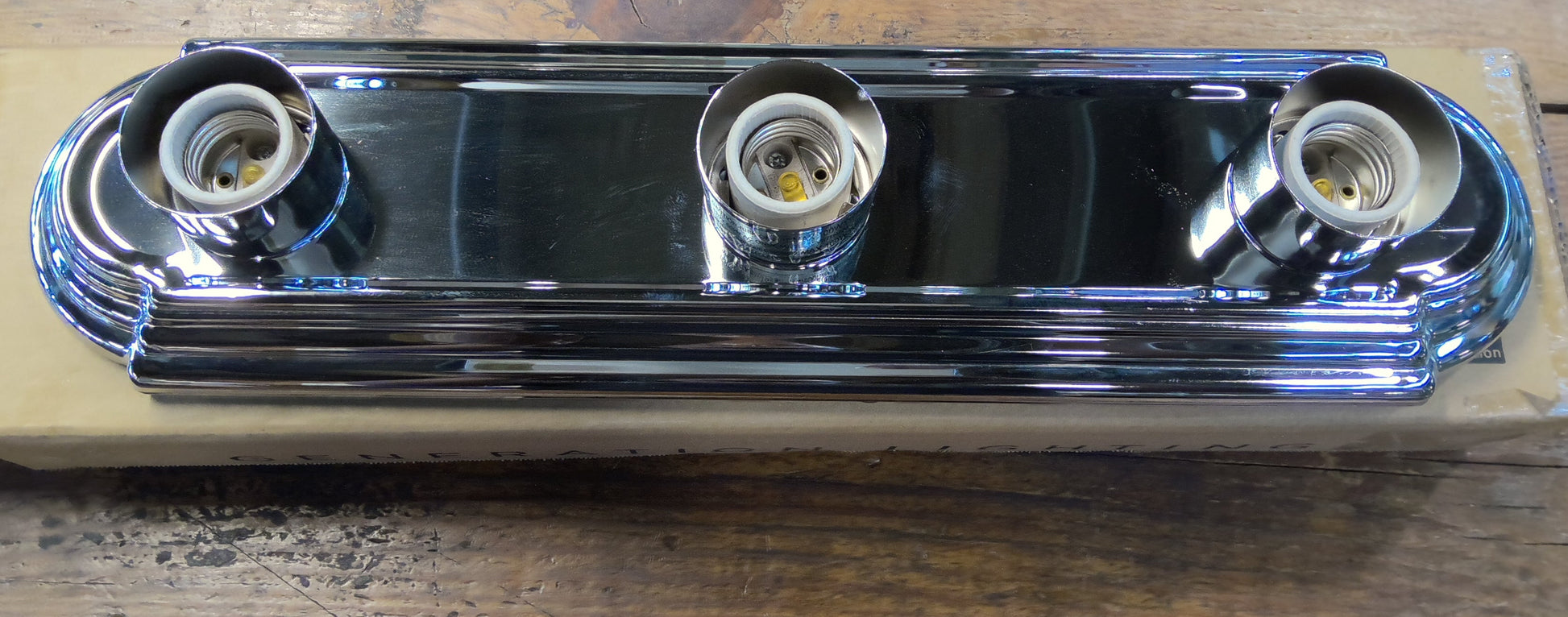 A three-light vanity light fixture with a polished chrome finish, resting on its cardboard packaging over a rustic wooden table.