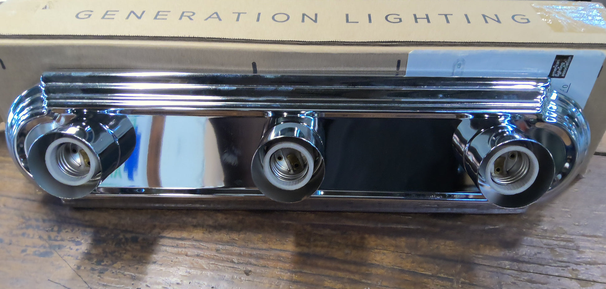 A three-light chrome wall sconce fixture with a polished metal finish, shown resting on a wooden workbench in front of its cardboard packaging box printed with 'GENERATION LIGHTING'.