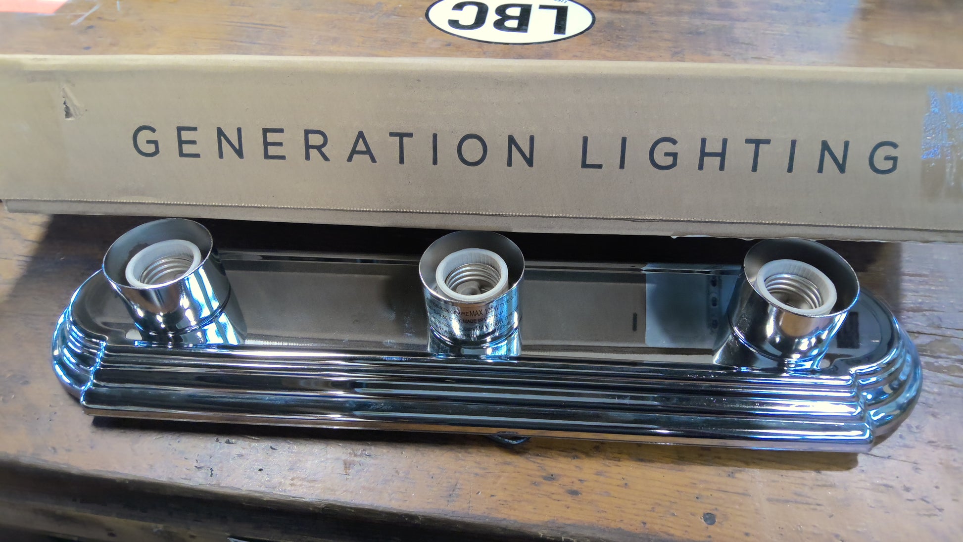 A three-light chrome wall sconce with ribbed decorative trim, shown in front of its cardboard box printed with 'GENERATION LIGHTING' and a small 'LBC' logo, resting on a wooden surface.