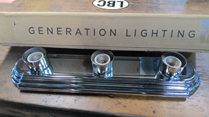 A three-light chrome wall sconce with ribbed decorative trim, shown in front of its cardboard box printed with 'GENERATION LIGHTING' and a small 'LBC' logo, resting on a wooden surface.