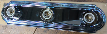 A chrome-plated metal automotive light bar with three circular bulb sockets, resting on a piece of cardboard over a rustic wooden table.