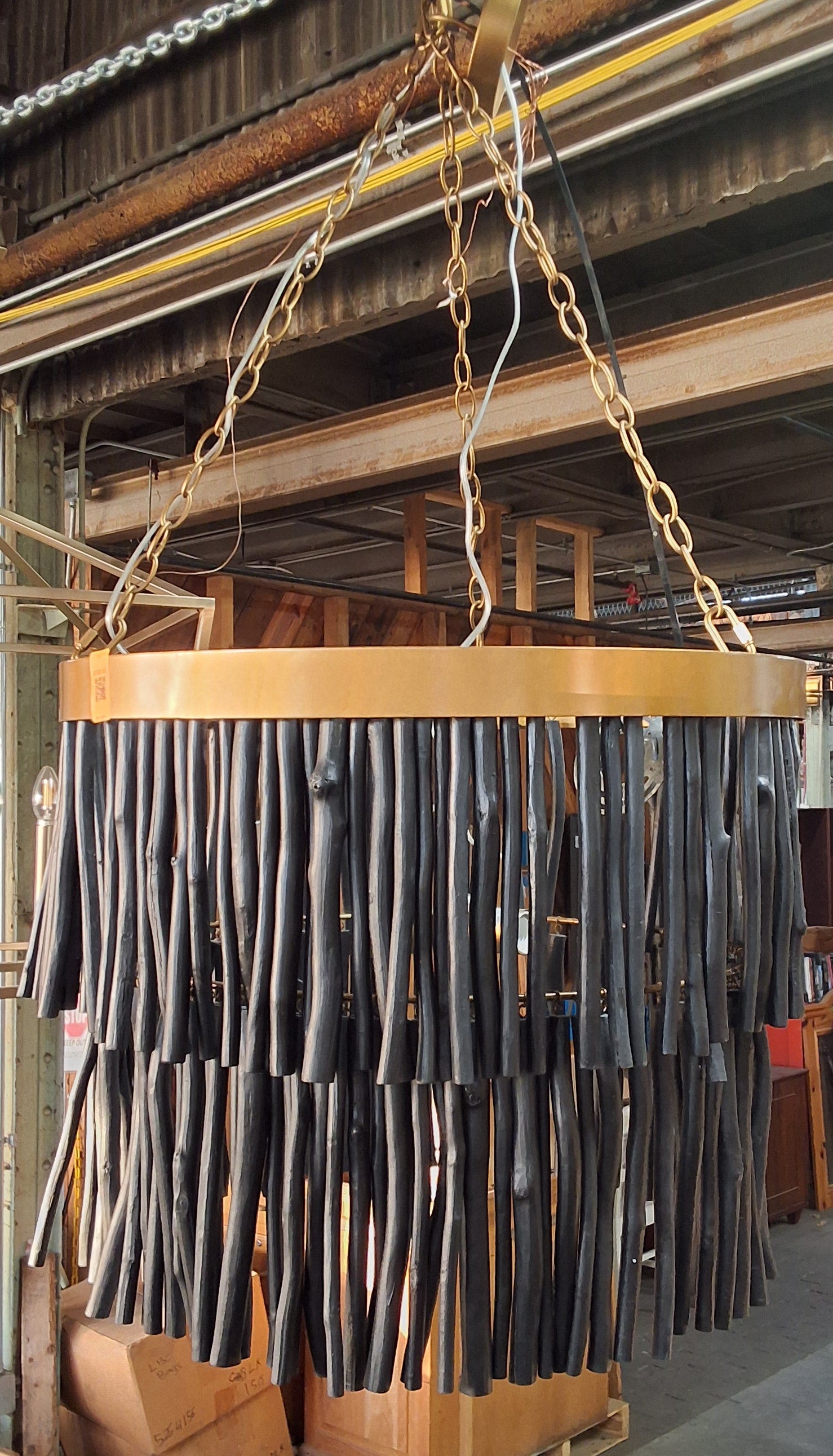 A large cylindrical hanging chandelier or pendant light made of many black rod-like elements suspended from a light wood ring, photographed inside a warehouse or showroom with chains and other furniture visible.
