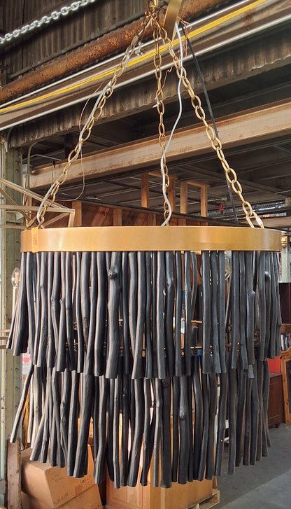 A large cylindrical hanging chandelier or pendant light made of many black rod-like elements suspended from a light wood ring, photographed inside a warehouse or showroom with chains and other furniture visible.