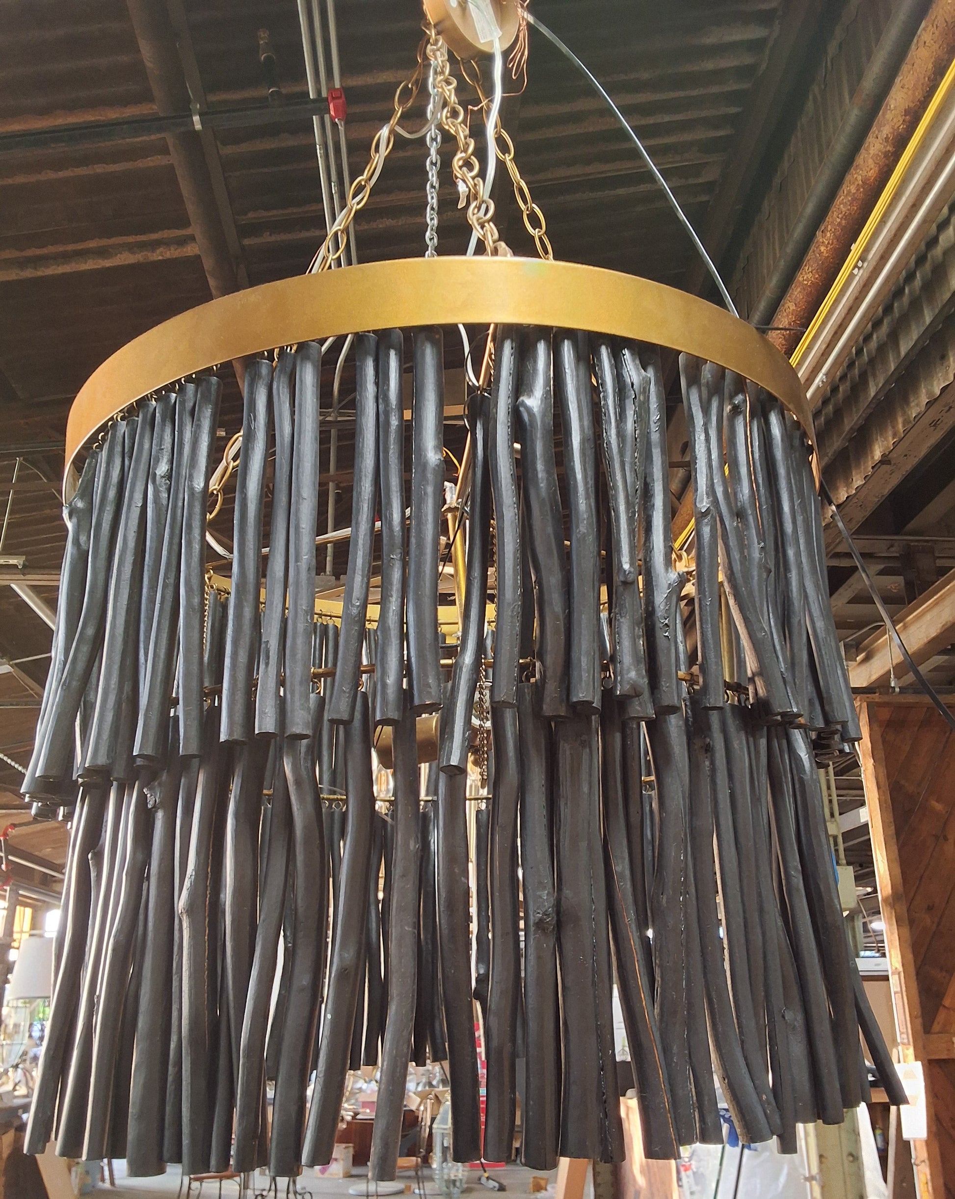 A large circular chandelier with a gold-toned metal frame and numerous dark, textured hanging rods, suspended by chains in an indoor workshop or warehouse setting.