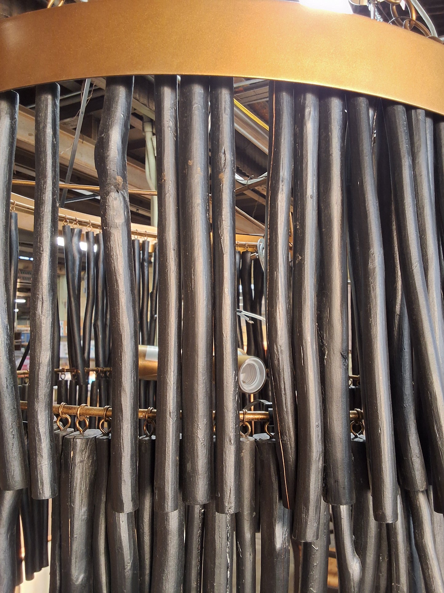 A close-up of a decorative wall hanging or chandelier made of many dark vertical rods or tubes, with a warm metallic gold curved band at the top and some brass-toned hardware visible behind it.
