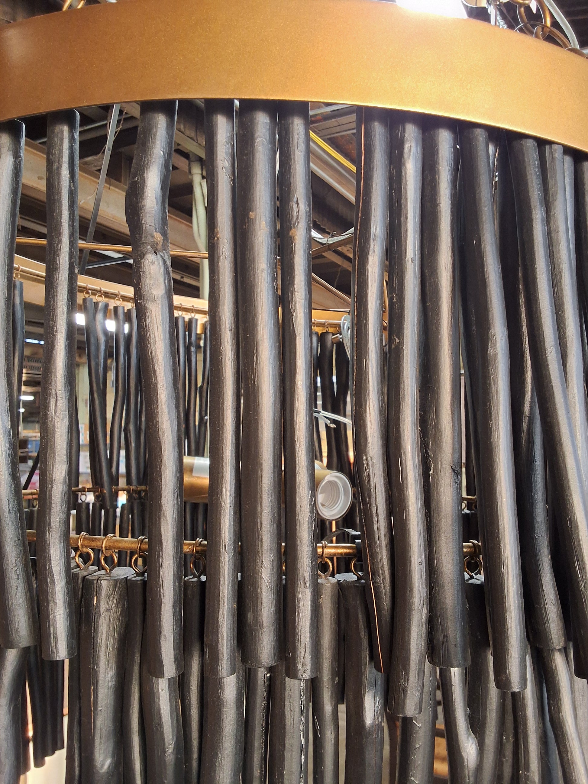 A close-up of a decorative wall hanging or chandelier made of many dark vertical rods or tubes, with a warm metallic gold curved band at the top and some brass-toned hardware visible behind it.