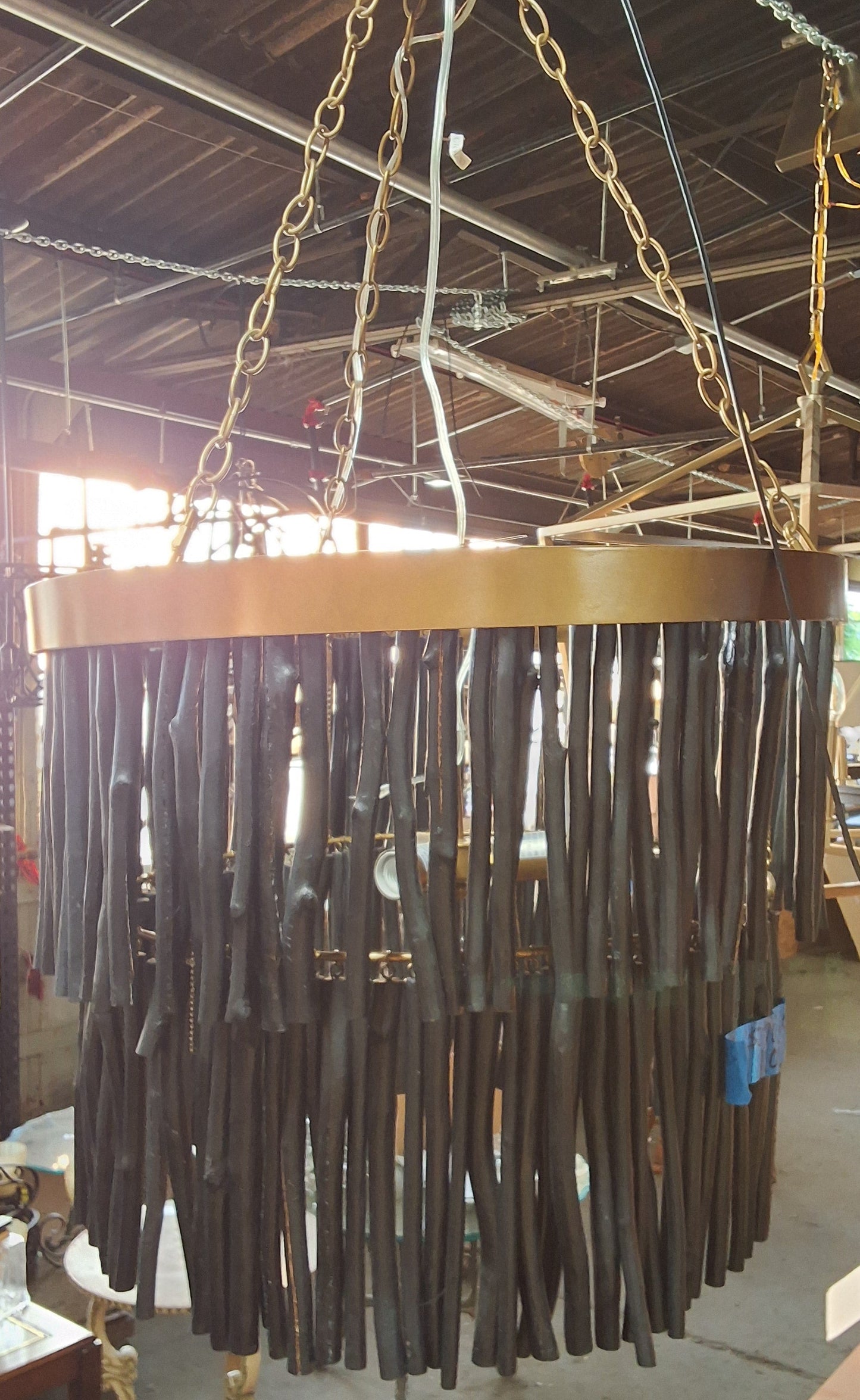 A large circular chandelier with a gold-toned metal frame and numerous dark brown hanging rods, suspended by chains in what appears to be a workshop or showroom.