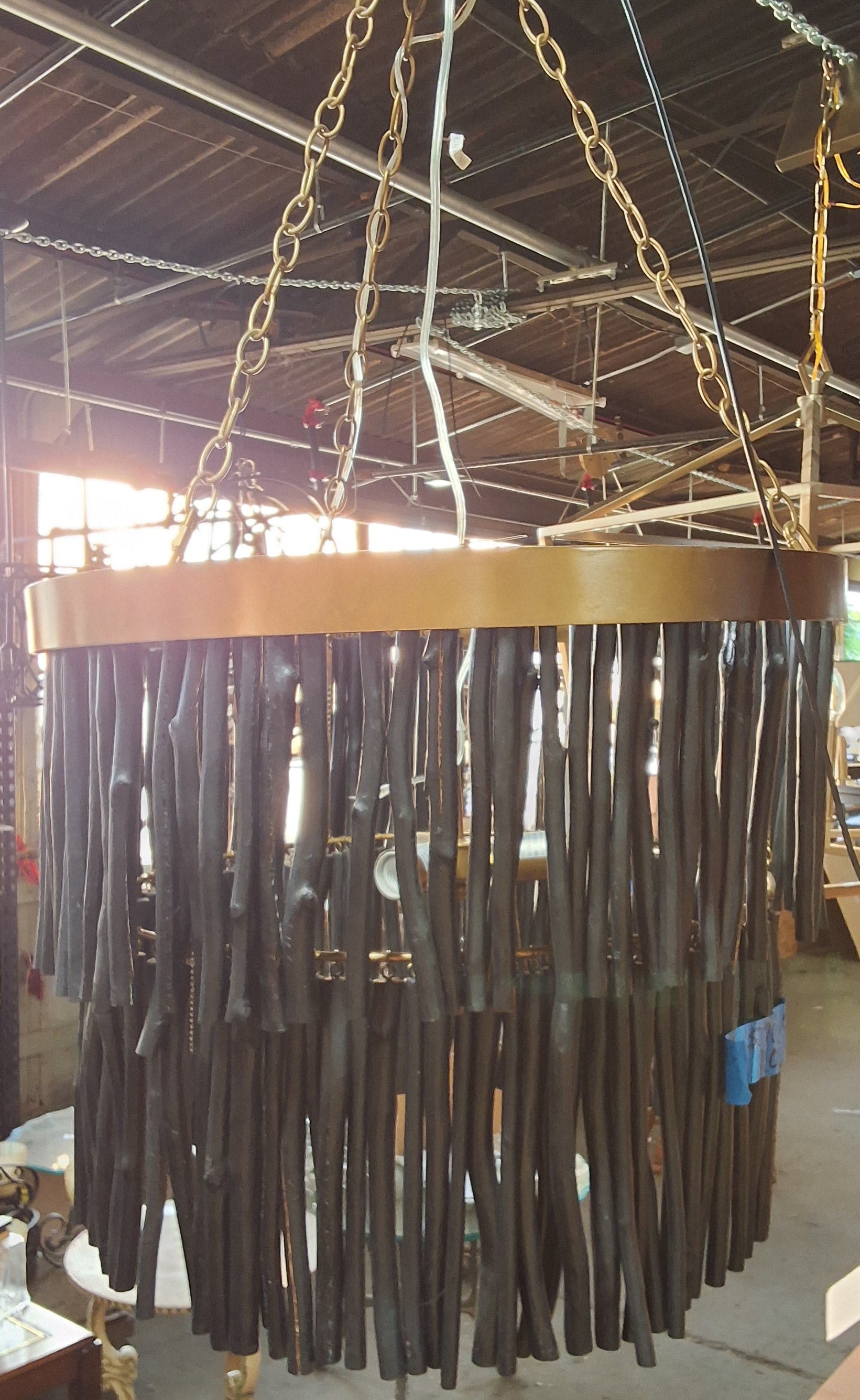 A large circular chandelier with a gold-toned metal frame and numerous dark brown hanging rods, suspended by chains in what appears to be a workshop or showroom.