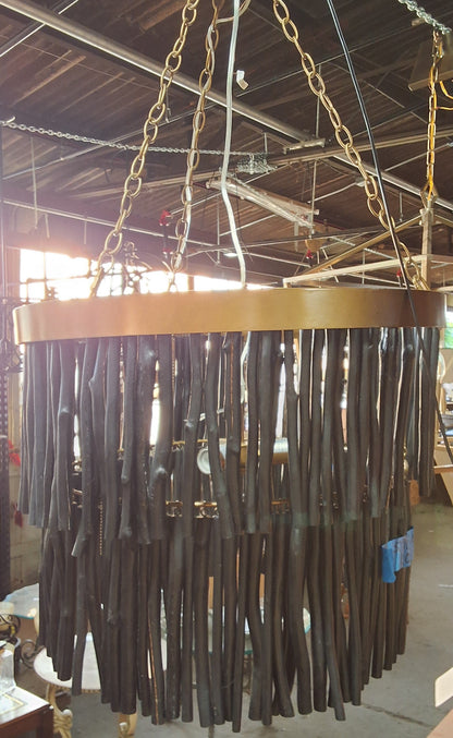 A large circular chandelier with a gold-toned metal frame and numerous dark brown hanging rods, suspended by chains in what appears to be a workshop or showroom.